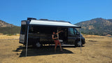 Enjoying an RV rental: A person poses by a dark grey camper van with extended awning, in a sunny, dry, mountainous landscape.