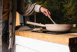 Cooking in a mobile RV rental kitchen. Person stirs a pan on the stovetop, green nature visible through the window.