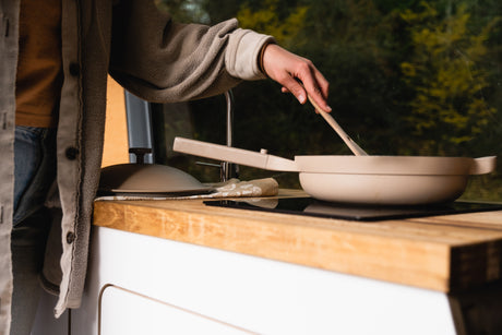 Cooking in a mobile RV rental kitchen. Person stirs a pan on the stovetop, green nature visible through the window.