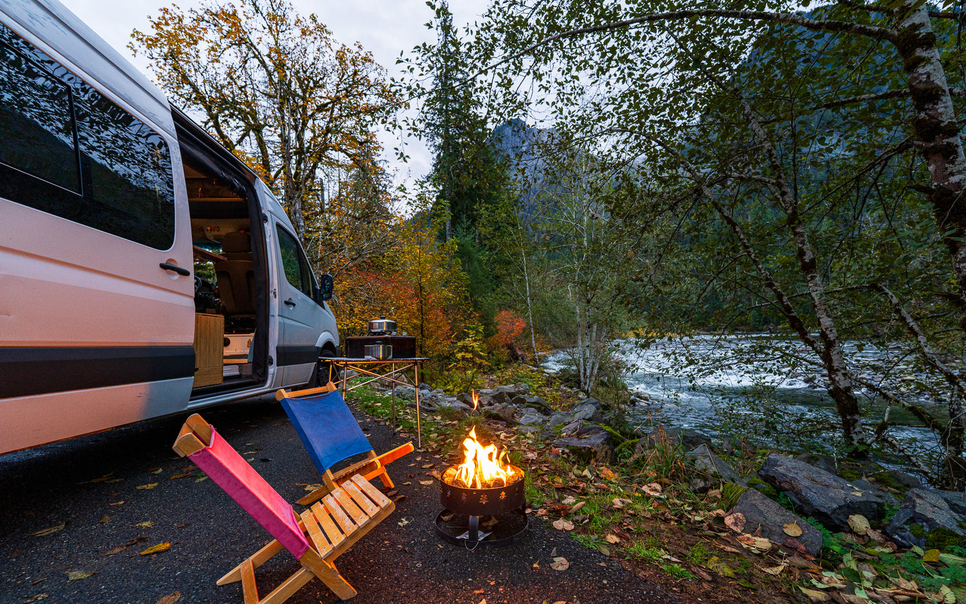 An rv rental camper van is parked by an autumn river with chairs, a crackling campfire, and a cooking setup.