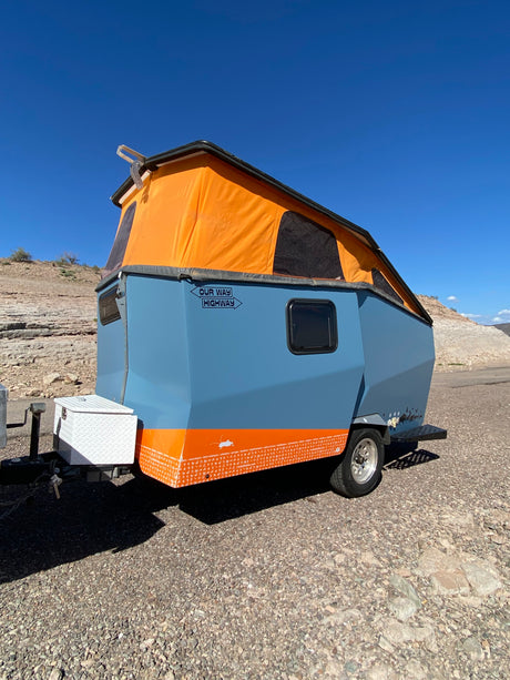 This unique blue & orange pop-top camper trailer, ideal for an RV rental, is parked in a rocky desert under a clear blue sky.