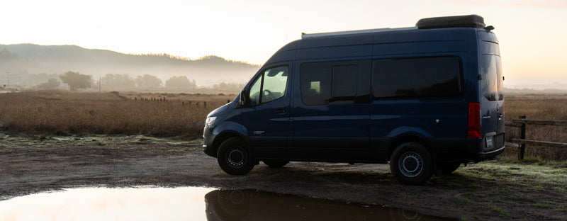 Blue RV rental van in a misty field at dawn, reflected in a puddle. — photo 3