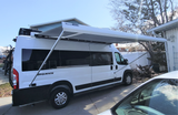 White Sequence camper van RV rental with a large Thule awning extended, parked in a sunny driveway.