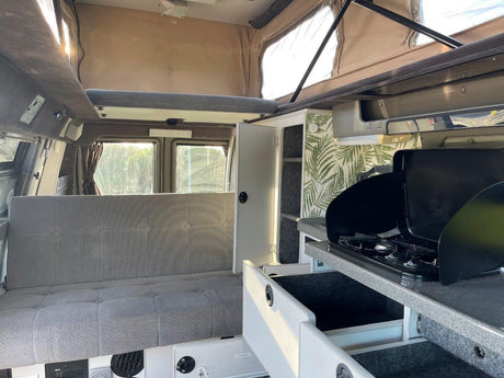 Inside an RV rental: view of the gray upholstered seating/bed area, kitchen with stove, and pop-top ceiling. Ready for adventure.