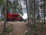 Red RV rental campervan parked at a scenic forest campsite with a picnic table, fire pit, and distant mountains.