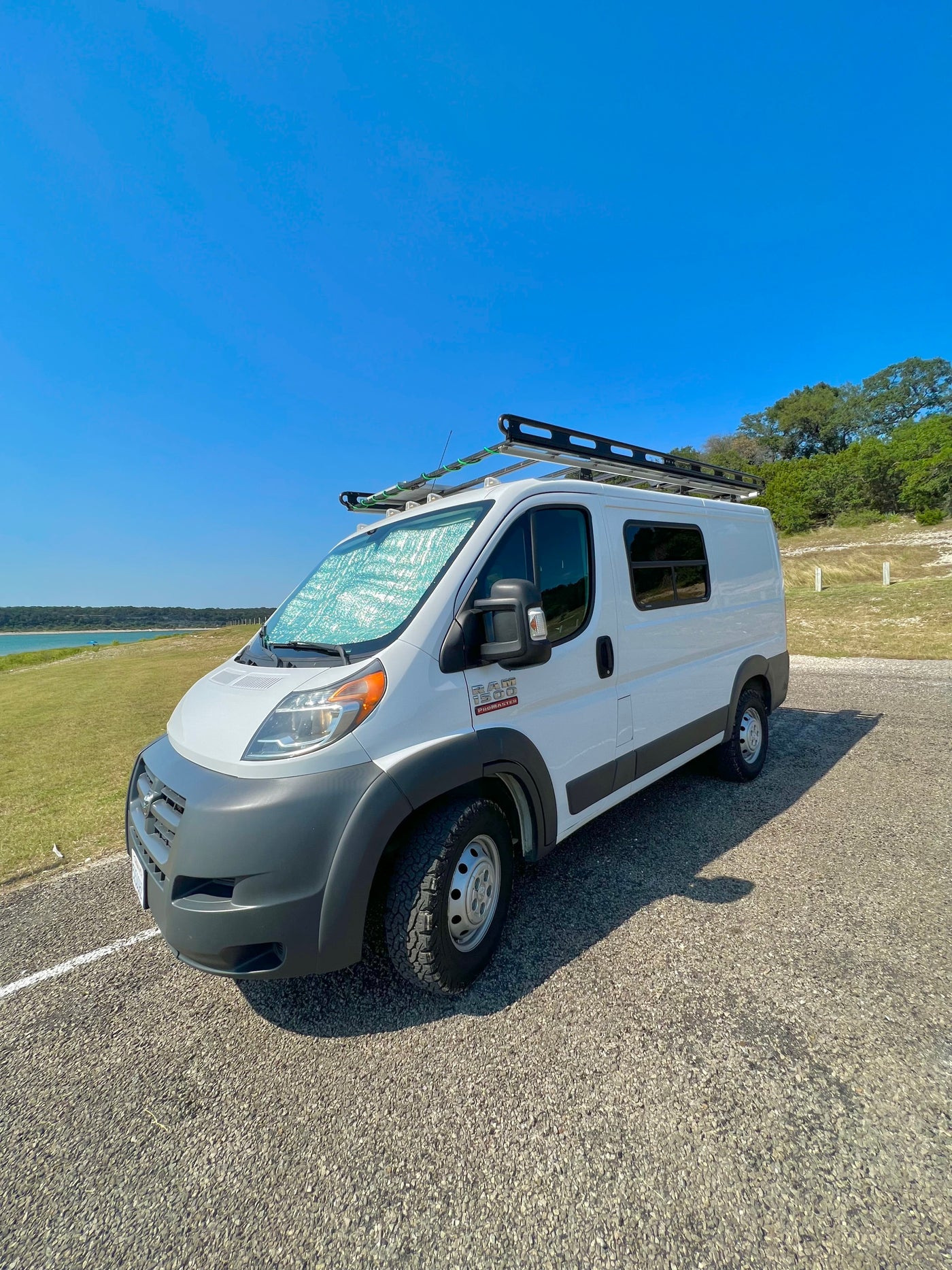 White Ram ProMaster rv rental van with roof rack, parked by a lake under a clear sky. Sunshade on windshield. — photo 16