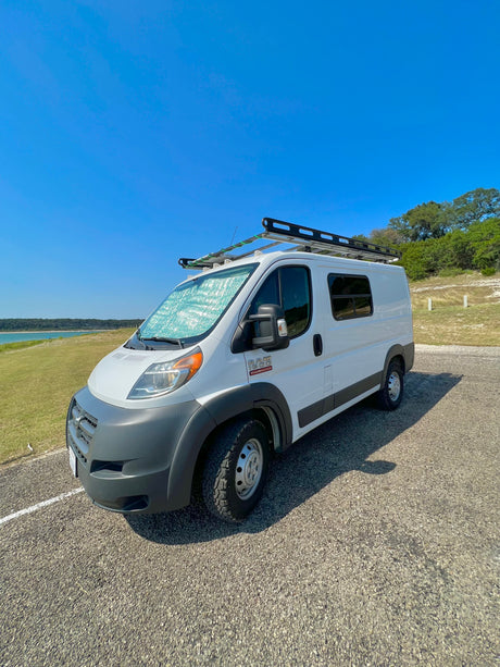 White Ram ProMaster rv rental van with roof rack, parked by a lake under a clear sky. Sunshade on windshield.