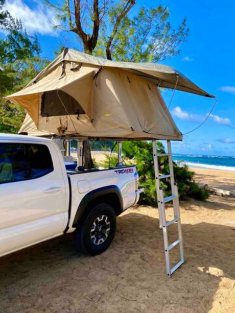 RV rental: White pickup truck with a tan rooftop tent & ladder set up on a beach. Trees & ocean visible behind.