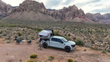 Adventure-ready RV rental pickup with rooftop tent and camp setup in a vast desert landscape with rugged mountains.