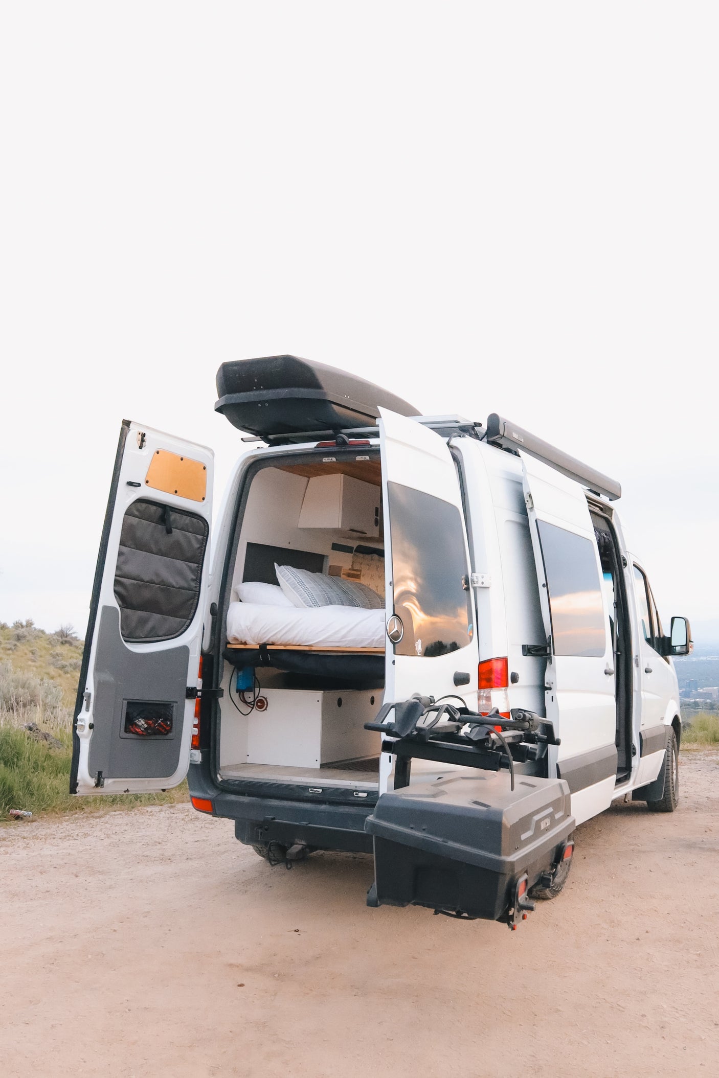Spacious RV rental camper van parked on a dirt road, rear doors open revealing bed, storage, and outdoor equipment.