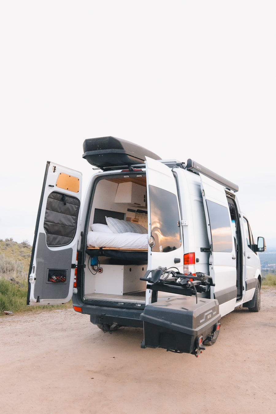 Spacious RV rental camper van parked on a dirt road, rear doors open revealing bed, storage, and outdoor equipment.