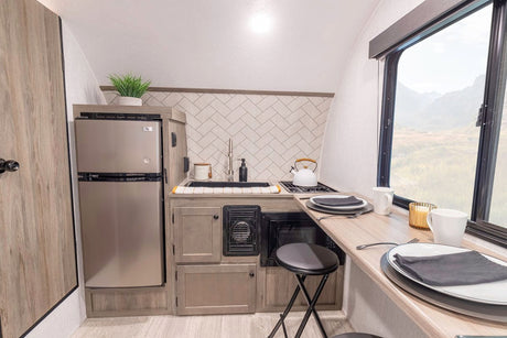 A modern RV rental kitchen and dining area features a stainless steel fridge, compact appliances, and a bar with a scenic window.