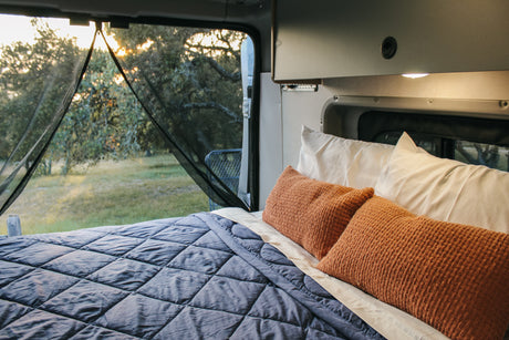 Cozy RV rental bedroom with blue comforter & orange pillows, offering serene nature views through a mesh screen.