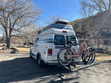 White Coachmen RV rental with two bikes on a rack, parked among bare trees and a hillside on a sunny day.