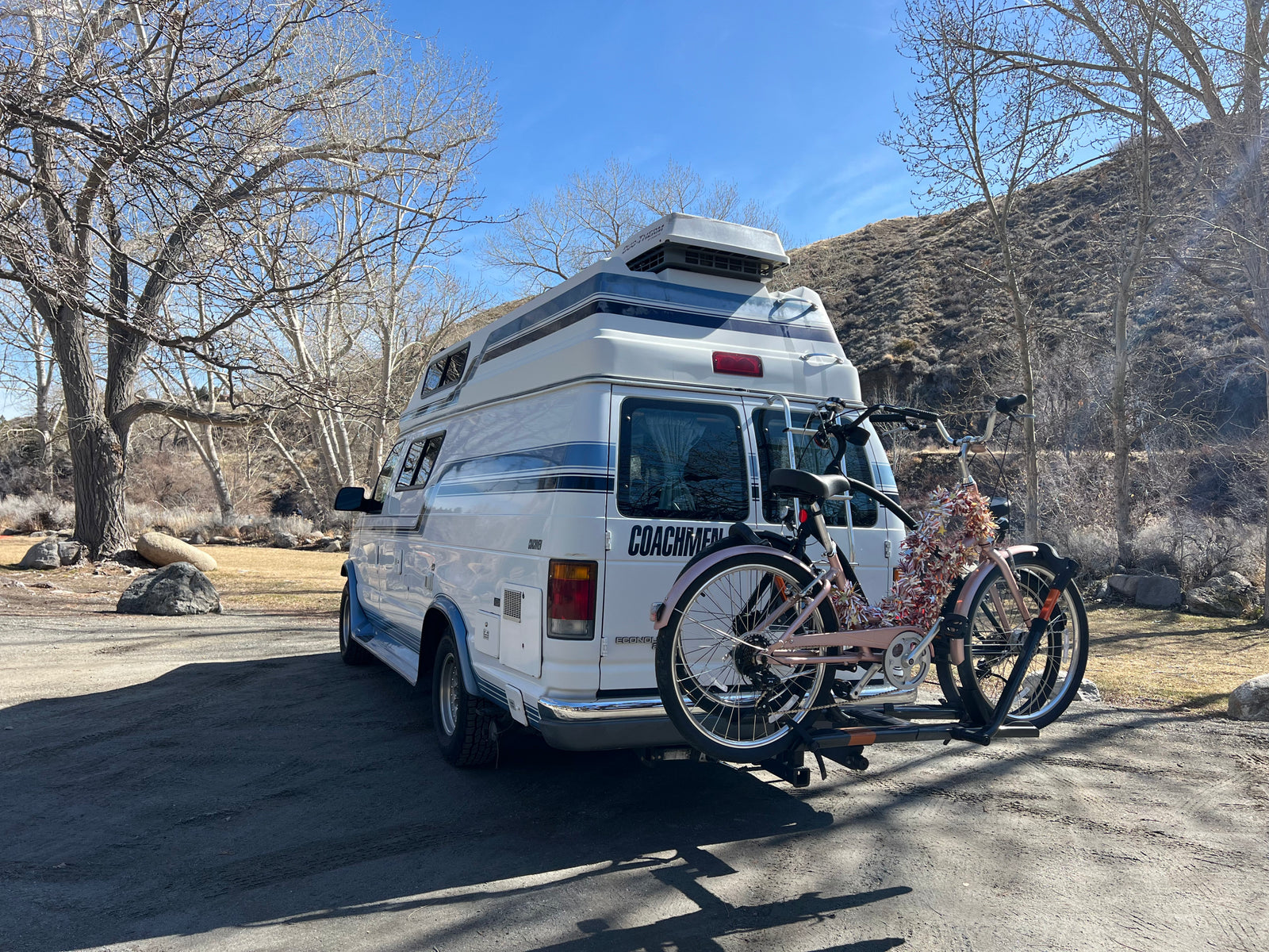 White Coachmen RV rental with two bikes on a rack, parked among bare trees and a hillside on a sunny day.