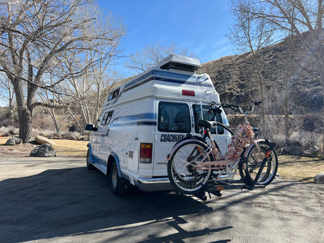 White Coachmen RV rental with two bikes on a rack, parked among bare trees and a hillside on a sunny day.