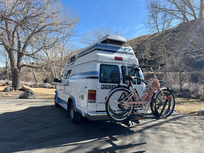 White Coachmen RV rental with two bikes on a rack, parked among bare trees and a hillside on a sunny day.
