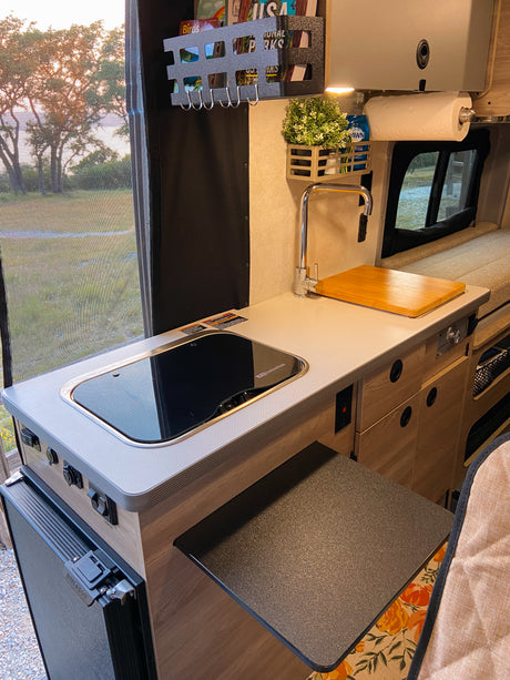 Inside a modern RV rental: a compact kitchen with sink, cooktop, mini-fridge, and clever storage, plus scenic window views.
