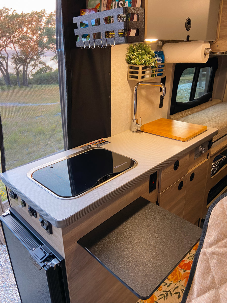 Inside a modern RV rental: a compact kitchen with sink, cooktop, mini-fridge, and clever storage, plus scenic window views.