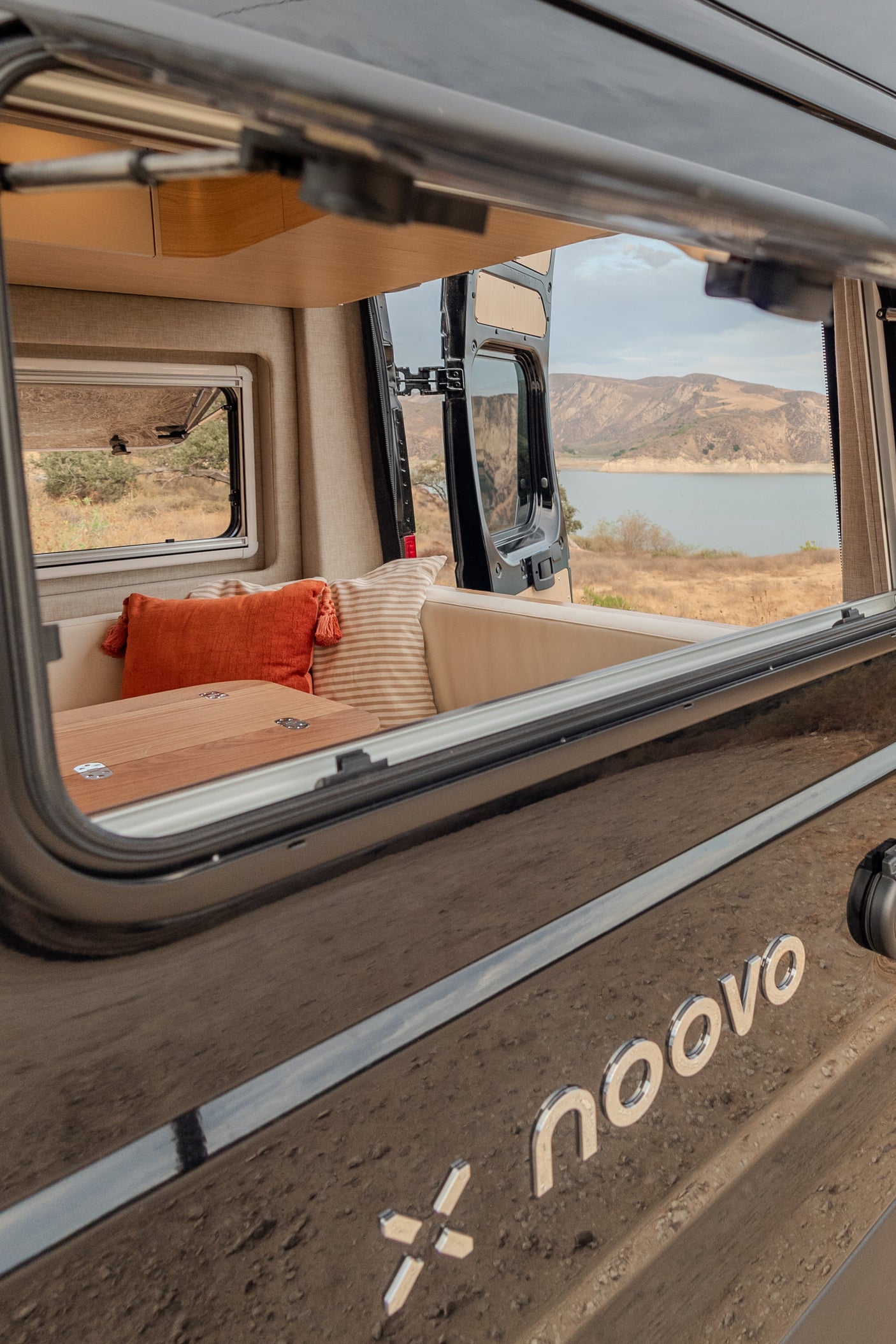 Enjoy your RV rental! The cozy camper van interior offers a stunning view of a calm lake and rolling hills, ideal for scenic travels.
