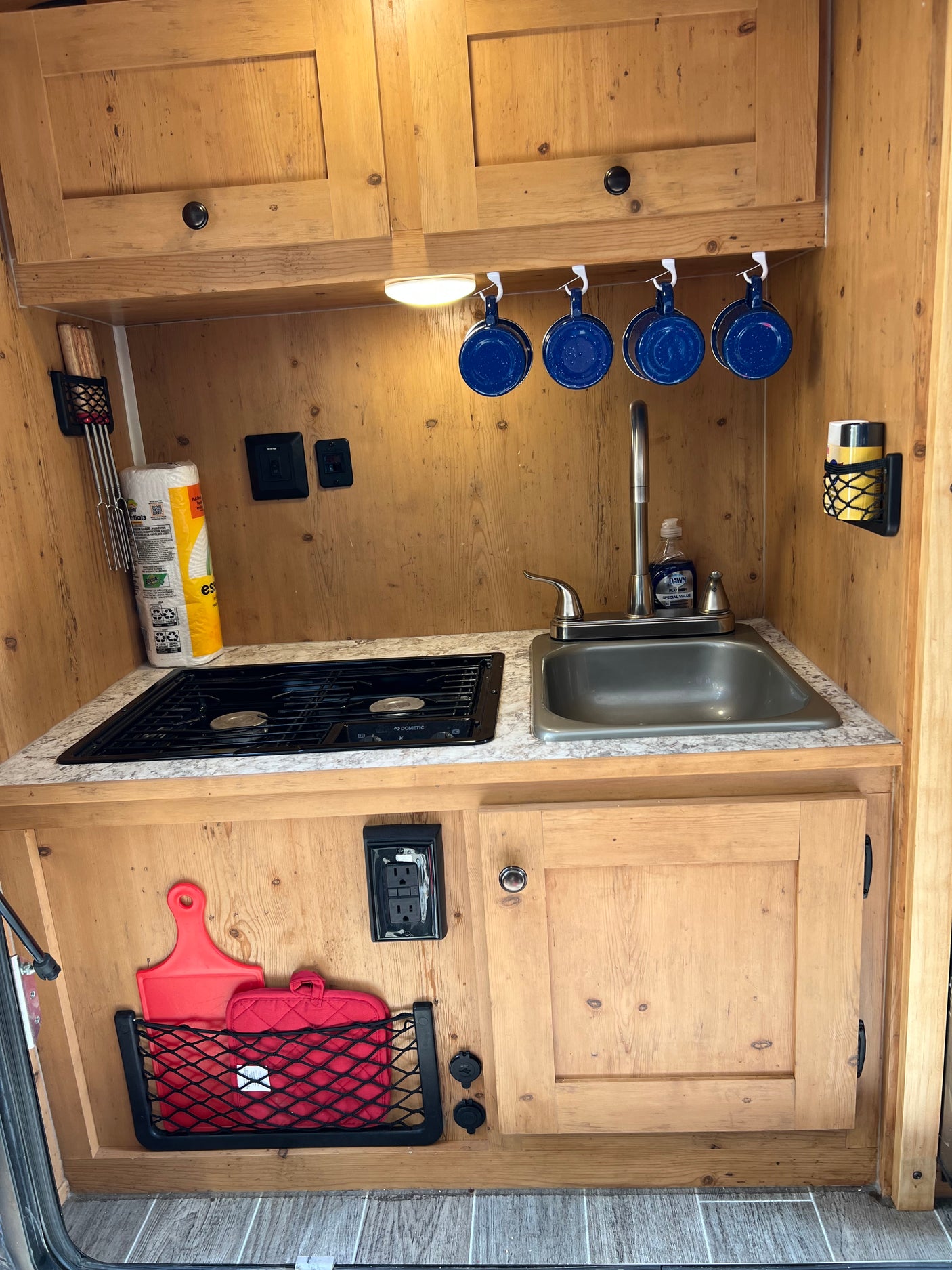 Explore this compact RV rental kitchen featuring a stove, sink, paper towels, and smart storage for mugs & utensils.
