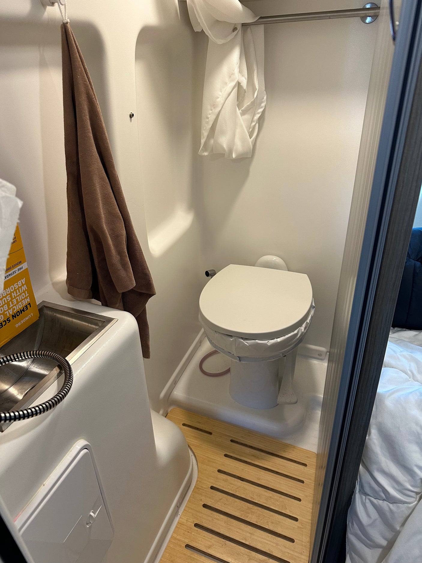 A bright, compact rv rental bathroom featuring a toilet with a bag liner, a sink with a shower sprayer, and a wooden floor mat.