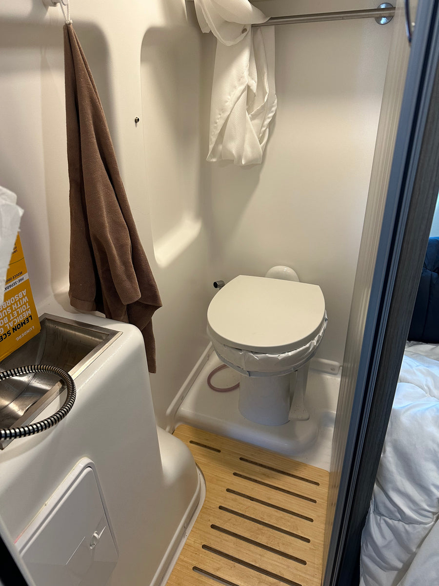 A bright, compact rv rental bathroom featuring a toilet with a bag liner, a sink with a shower sprayer, and a wooden floor mat.
