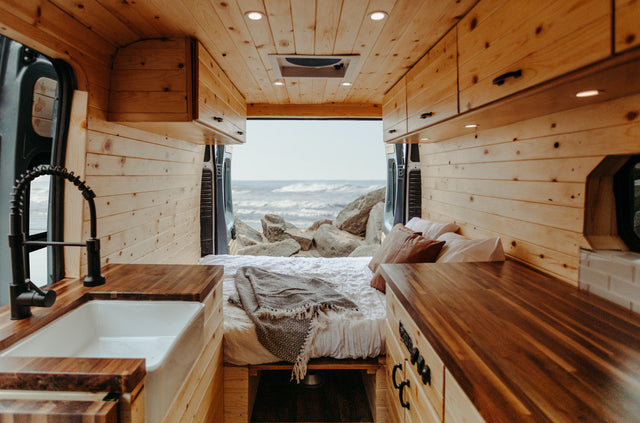 Inside a custom wood-paneled rv rental camper van. Features a kitchen, comfortable bed, and scenic ocean view for mobile living.