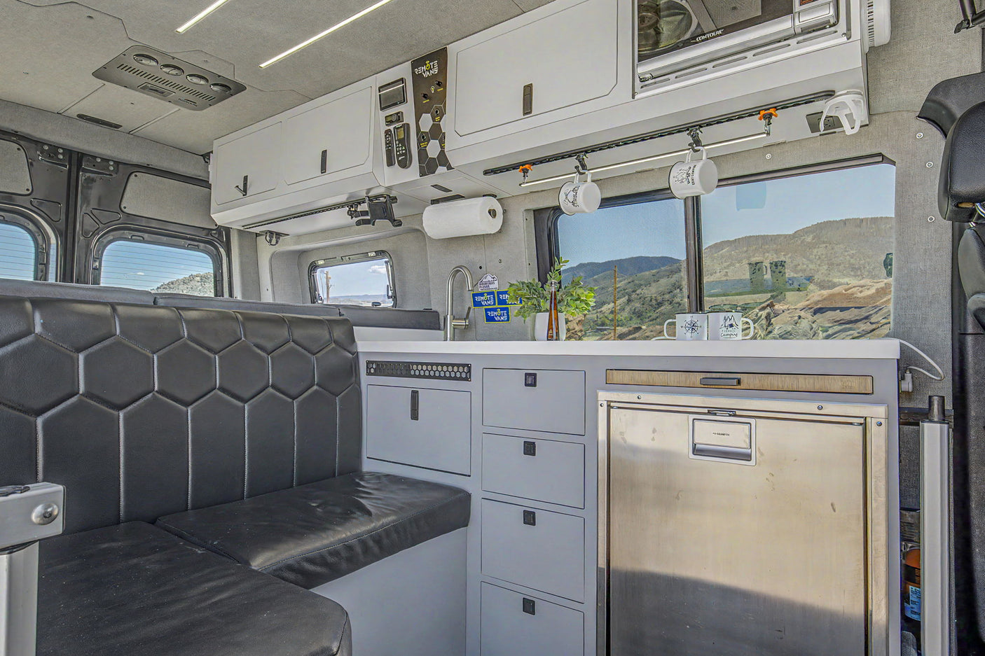Custom RV rental interior: Camper van kitchen with built-in fridge, hexagonal seating, and mountain views outside.