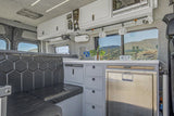 Custom RV rental interior: Camper van kitchen with built-in fridge, hexagonal seating, and mountain views outside.