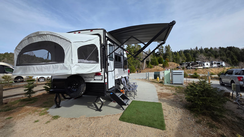An RV rental, a travel trailer with a pop-out bed and awning, is set up at a scenic campsite among trees. — photo 13