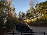 Black pop-top RV rental van on a gravel drive, nestled among fall trees with a golden sunset glowing through. Ideal for camping.