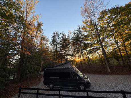 Black pop-top RV rental van on a gravel drive, nestled among fall trees with a golden sunset glowing through. Ideal for camping.