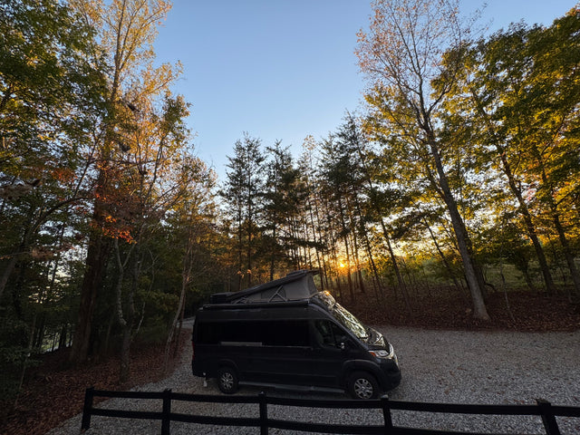 Black pop-top RV rental van on a gravel drive, nestled among fall trees with a golden sunset glowing through. Ideal for camping.