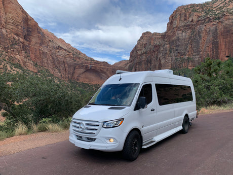 White Mercedes-Benz Sprinter RV rental parked amidst towering red rock canyon walls and lush green foliage.