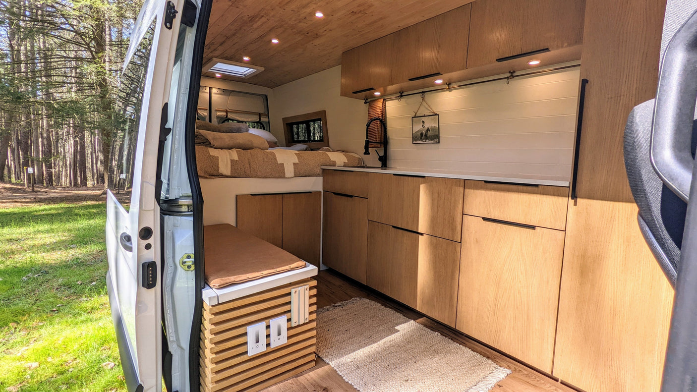 Spacious camper van interior with an open door revealing a forest view. Bed, kitchen, and seating are ready for RV rental.