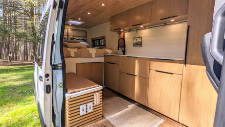 Spacious camper van interior with an open door revealing a forest view. Bed, kitchen, and seating are ready for RV rental.