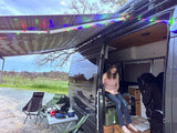 This RV rental offers comfort: a woman relaxes inside a camper van with an awning, colorful lights, & outdoor cooking in nature.