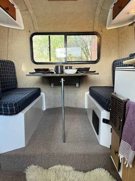 Compact RV rental interior: cozy dining area with plaid cushions, table, and window views.