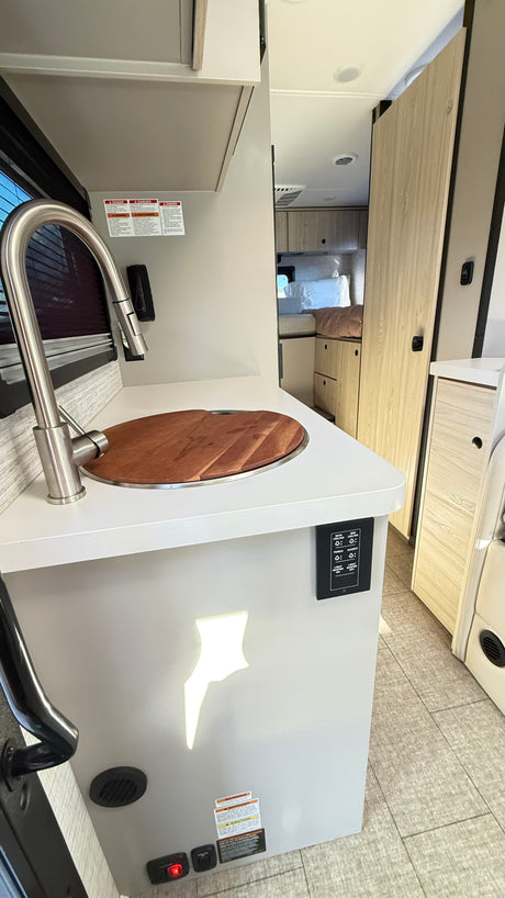 A bright, modern RV rental interior with a kitchen featuring a pull-down faucet, wooden sink cover, and controls. Beyond, a bed.