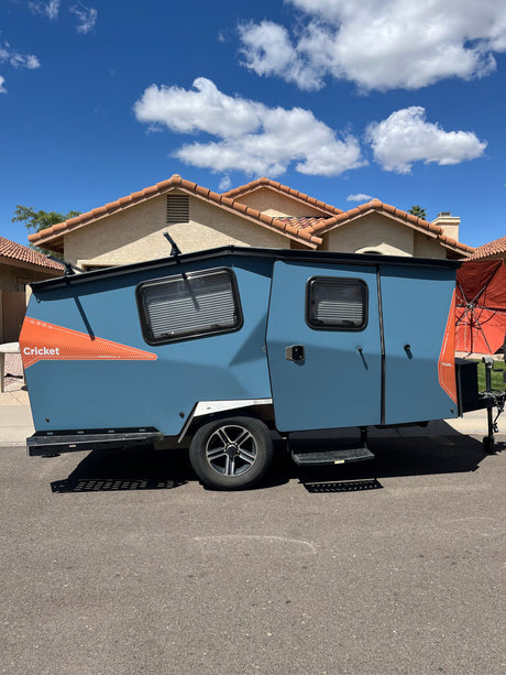 Blue Cricket travel trailer, a unique RV rental option, parked on asphalt. Ideal for outdoor adventures under clear skies.