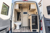 Compact RV rental interior view from rear: includes toilet, kitchen with sink/microwave, and wood-look floor.