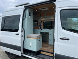 Explore this modern camper van RV rental interior! Features a custom light blue kitchenette, cozy seating, and a warm wooden ceiling.