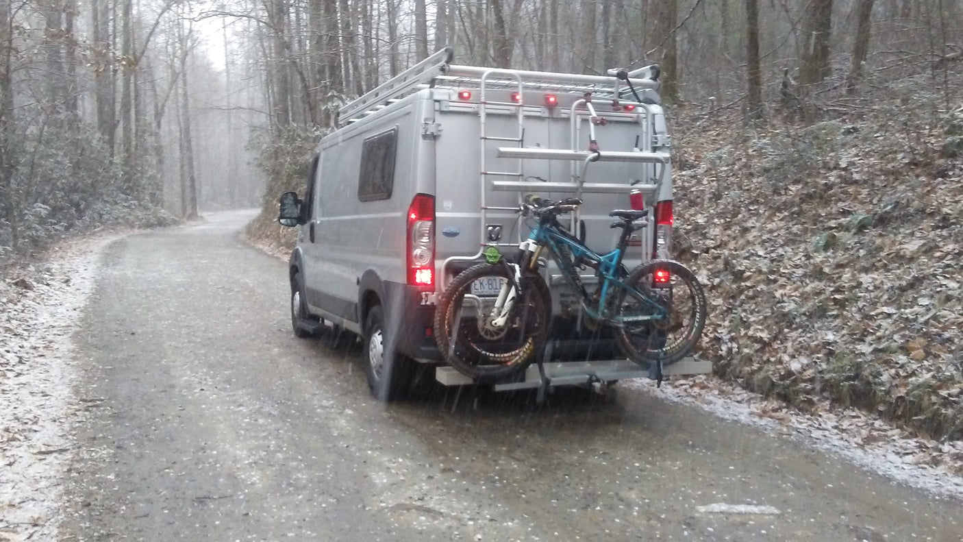 Gray RV rental van with mountain bikes on a snowy forest road. Ideal for winter adventures.