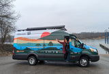 A smiling woman stands in a vibrant RV rental van, decorated with a landscape mural, by a river and dock. Great for travel.