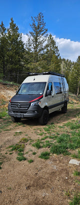 Light-colored Mercedes Sprinter camper van, with a roof rack and ladder, ideal for an RV rental, parked in a dirt forest clearing.