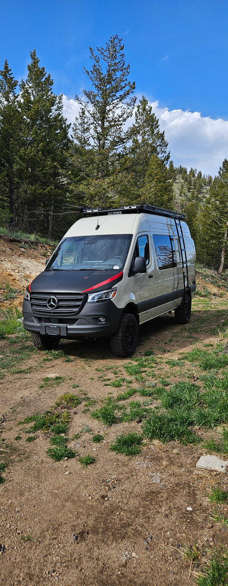 Light-colored Mercedes Sprinter camper van, with a roof rack and ladder, ideal for an RV rental, parked in a dirt forest clearing.