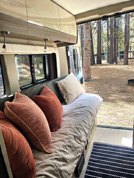 Cozy RV rental interior with plush cushions and a view of tall forest trees at a peaceful campsite.