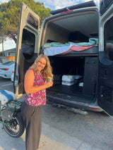 Smiling woman enjoys her RV rental camper van, revealing a cozy bed, portable toilet, and e-bike.
