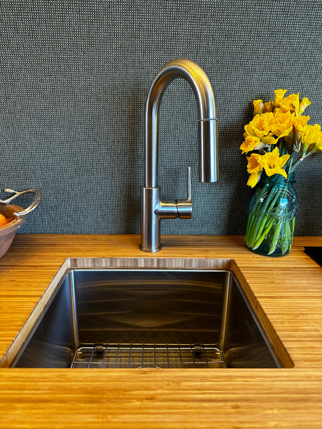 Sleek RV rental kitchen: bamboo counter, stainless sink with pull-down faucet, dark textured backsplash, and yellow flowers.
